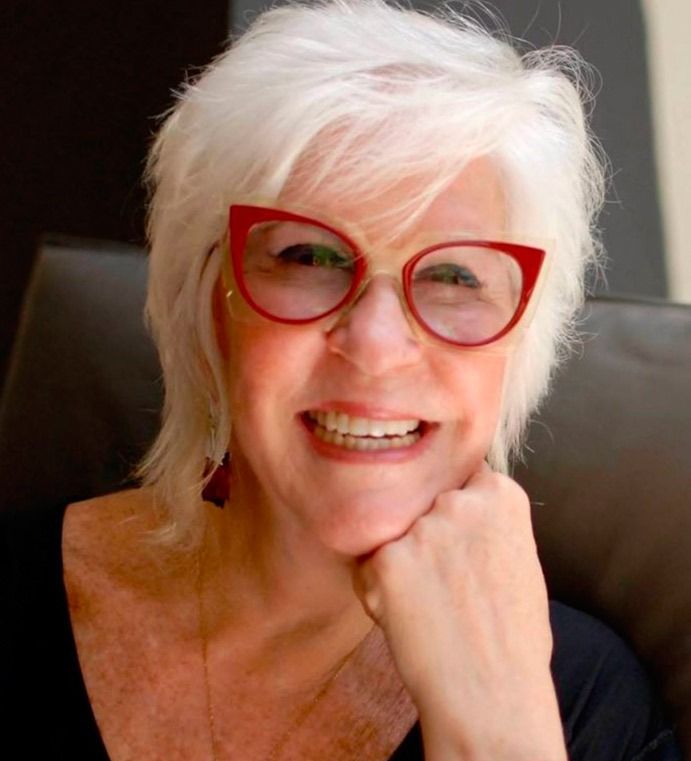 Una mujer con cabello blanco y gafas rojas está sonriendo.