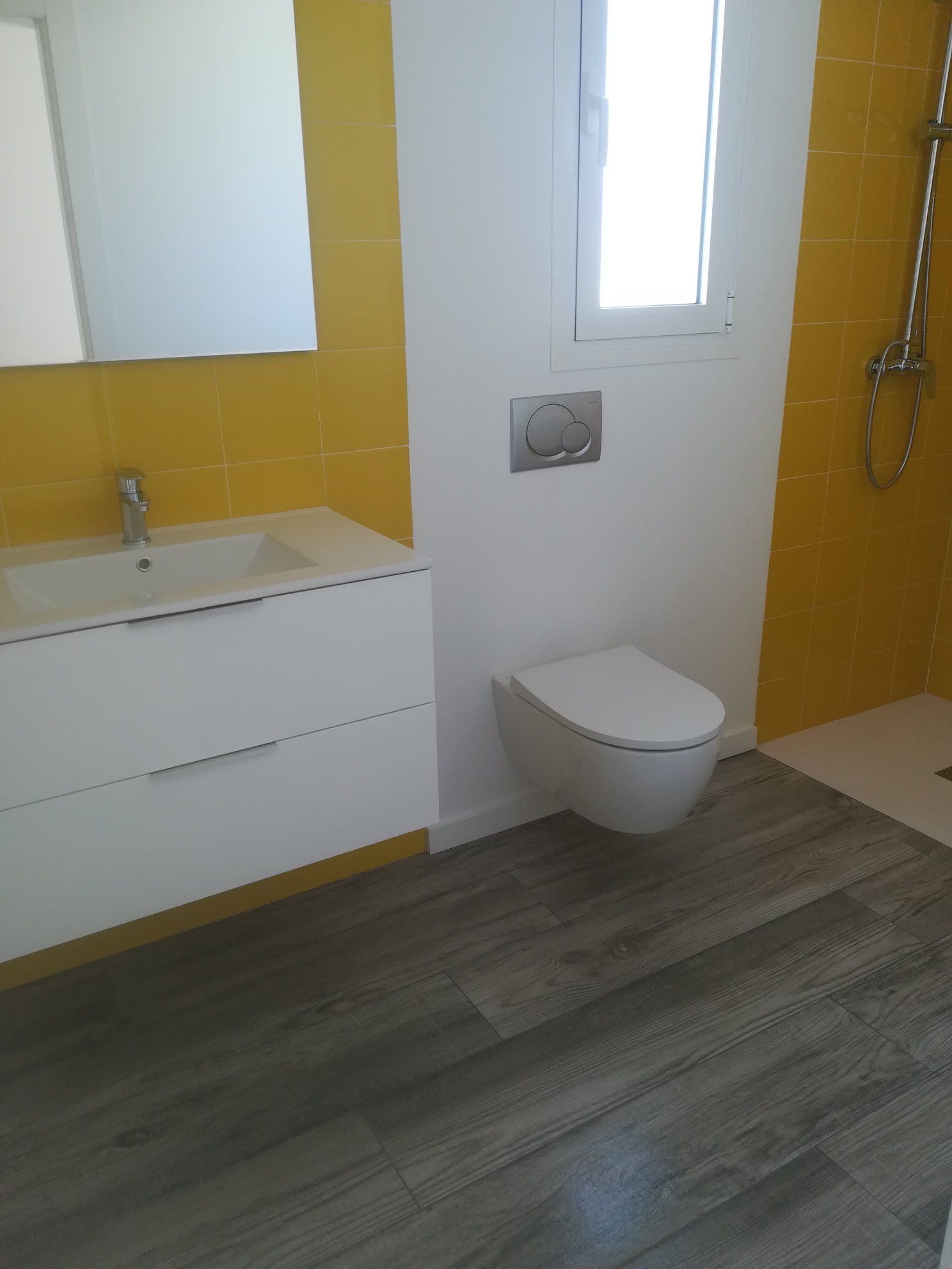 Baño con paredes revestidas de azulejos amarillos, lavabo blanco e inodoro suspendido; suelo con aspecto de madera.