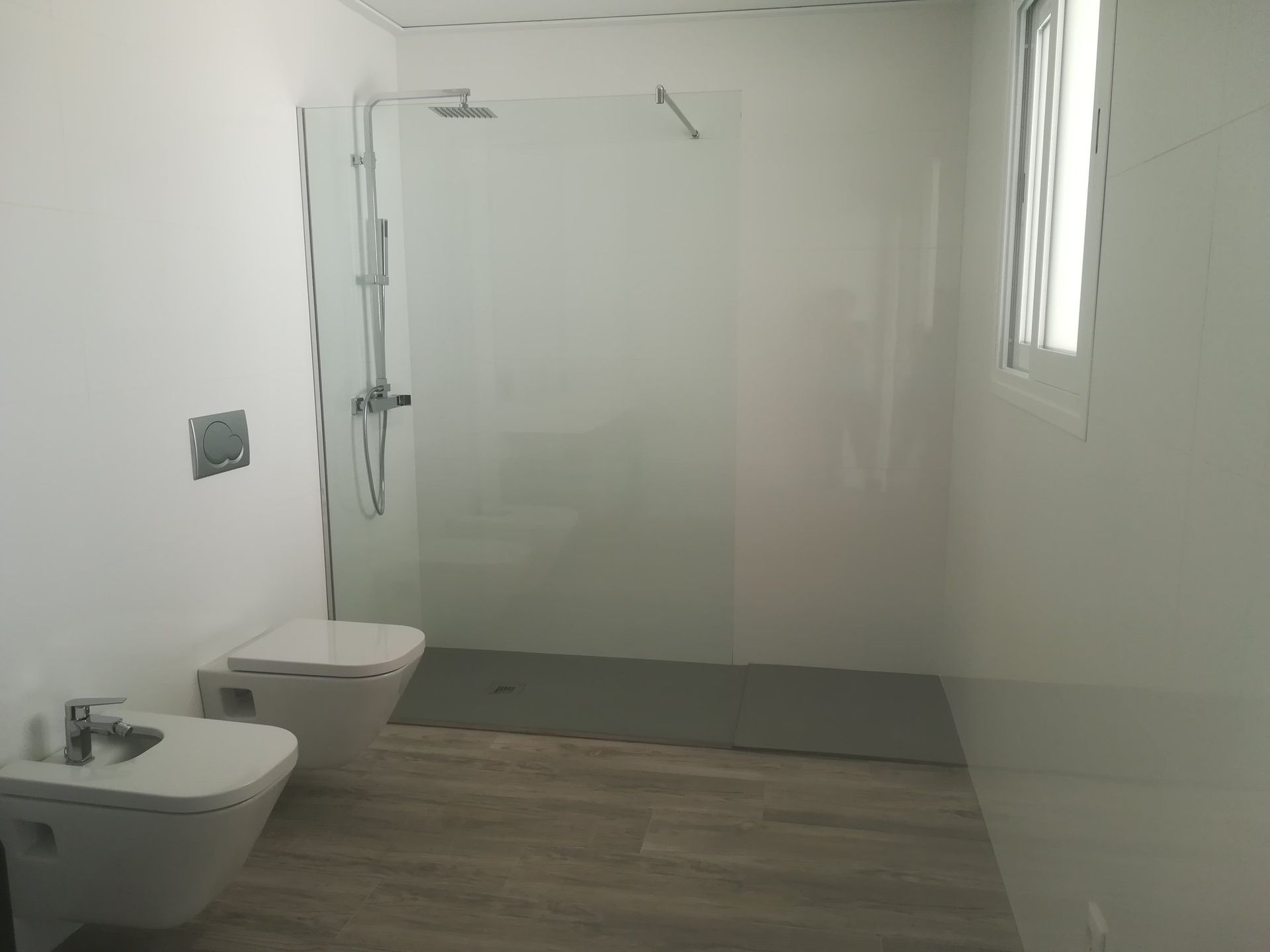 Baño moderno blanco con ducha, inodoro y bidé. Suelo de madera, gran mampara de ducha de cristal.