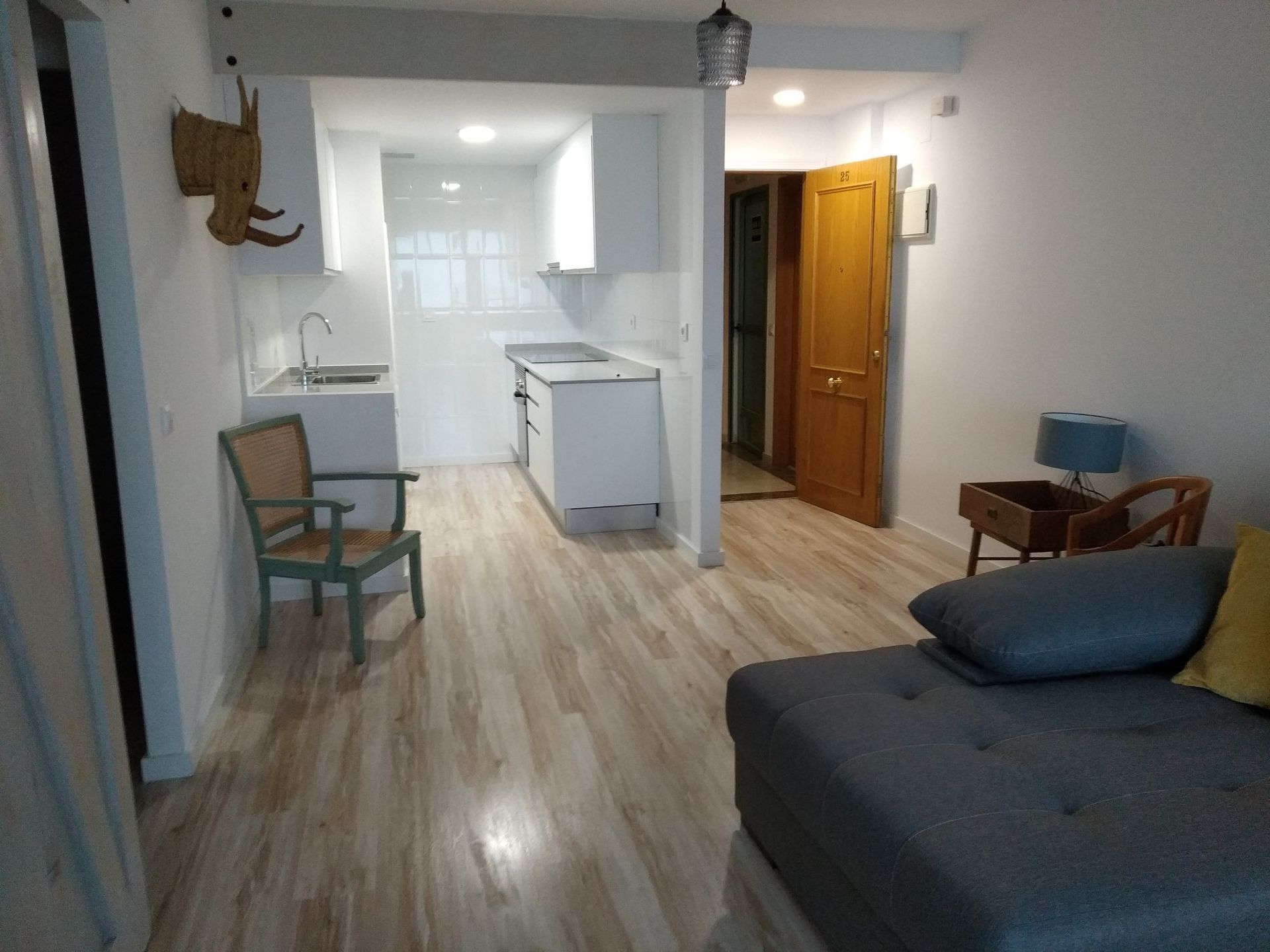 Espacio luminoso y diáfano con suelos de madera clara, cocina y sofá seccional gris.