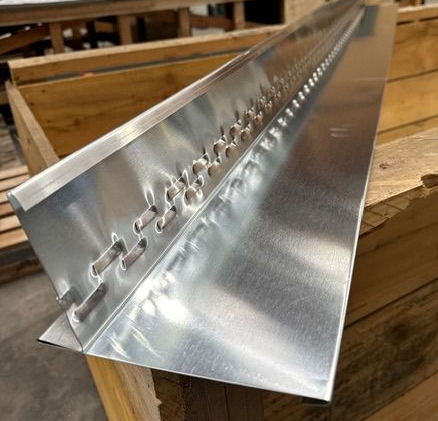 Une longue pièce de métal courbée, percée de trous régulièrement espacés, reposant sur des palettes en bois.