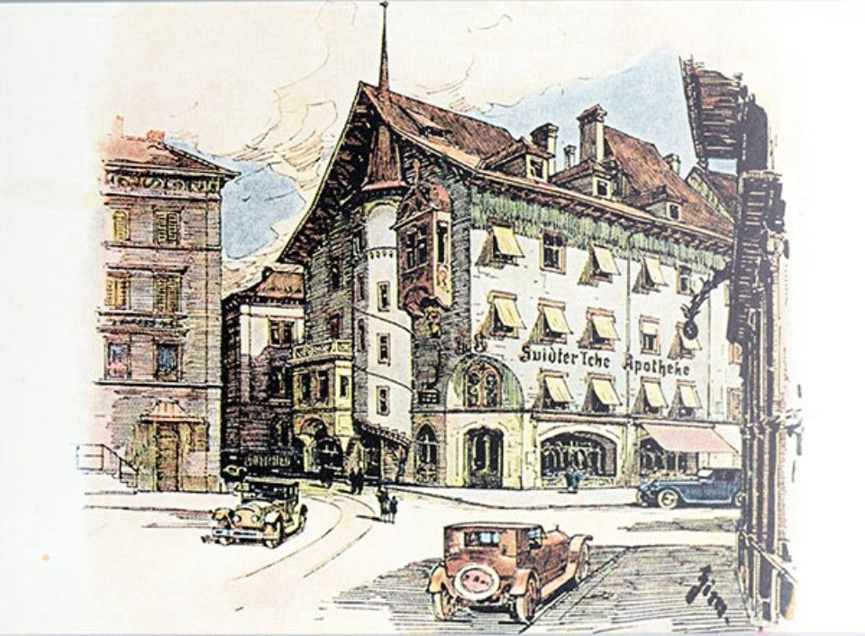 Aquarellillustration eines Gebäudes, der „Goldener Lobe Apotheke“, mit Autos auf einer Strasse. Braune und beige Farbtöne.