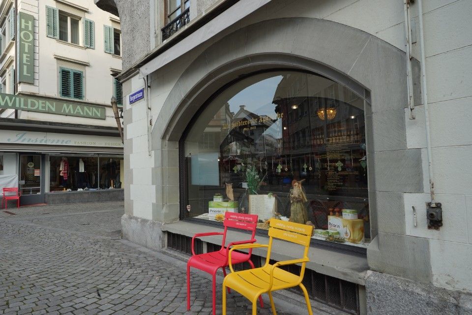 Ein Laden mit einem grossen Bogenfenster spiegelt das Kopfsteinpflaster einer Strasse wider. Draussen stehen rote und gelbe Stühle. Schild: „Hotel Wilden Mann“.