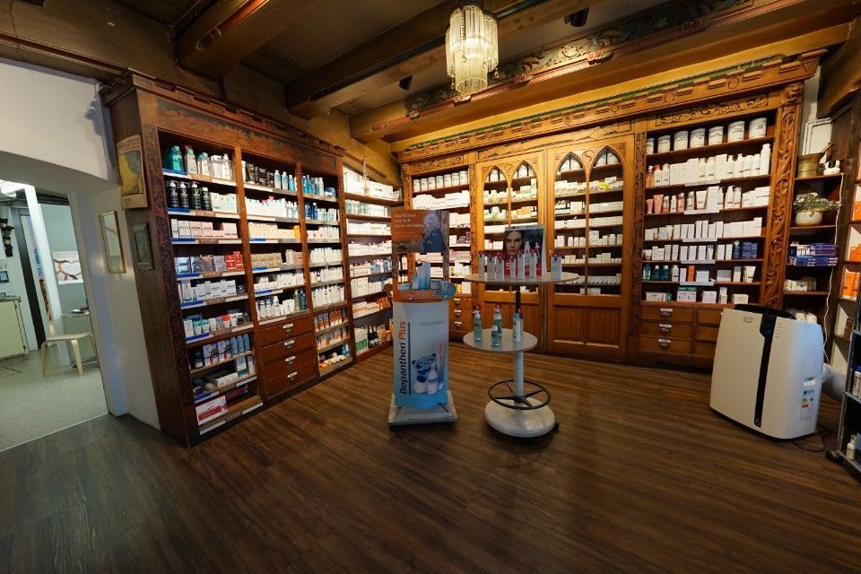 Innenansicht einer Apotheke mit hölzernen Regalen voller Produkte; einem zentralen Auslagebereich und dem Boden.