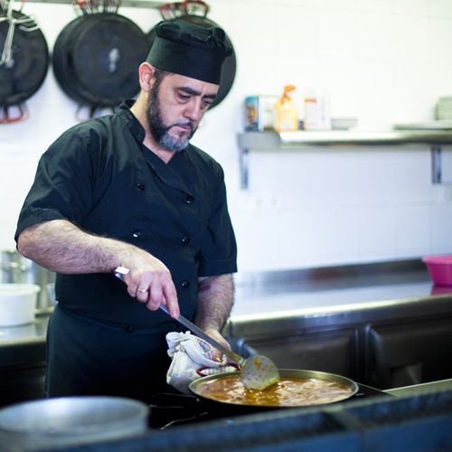 Chef con uniforme negro revolviendo comida en una sartén sobre una estufa, en una cocina comercial.