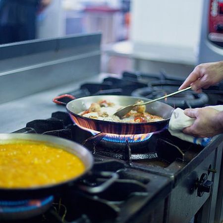 Manos revolviendo comida en sartenes sobre una estufa de gas, con una olla de sopa amarilla hirviendo a fuego lento cerca.