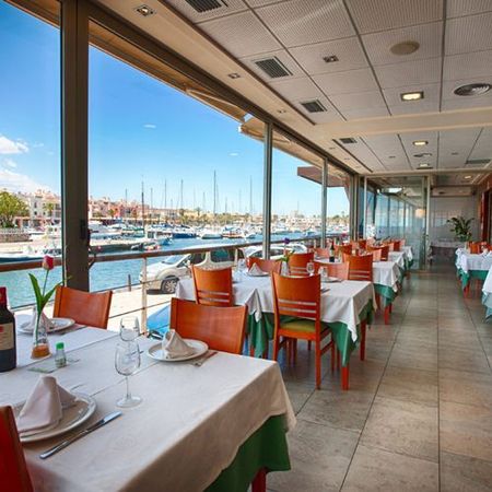 Comedor de restaurante junto al puerto con manteles blancos, sillas naranjas y vistas al puerto deportivo a través de grandes ventanales.