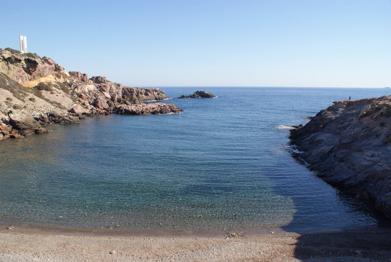 Cala rocosa con aguas tranquilas y azules. Un faro se alza sobre un acantilado a lo lejos.