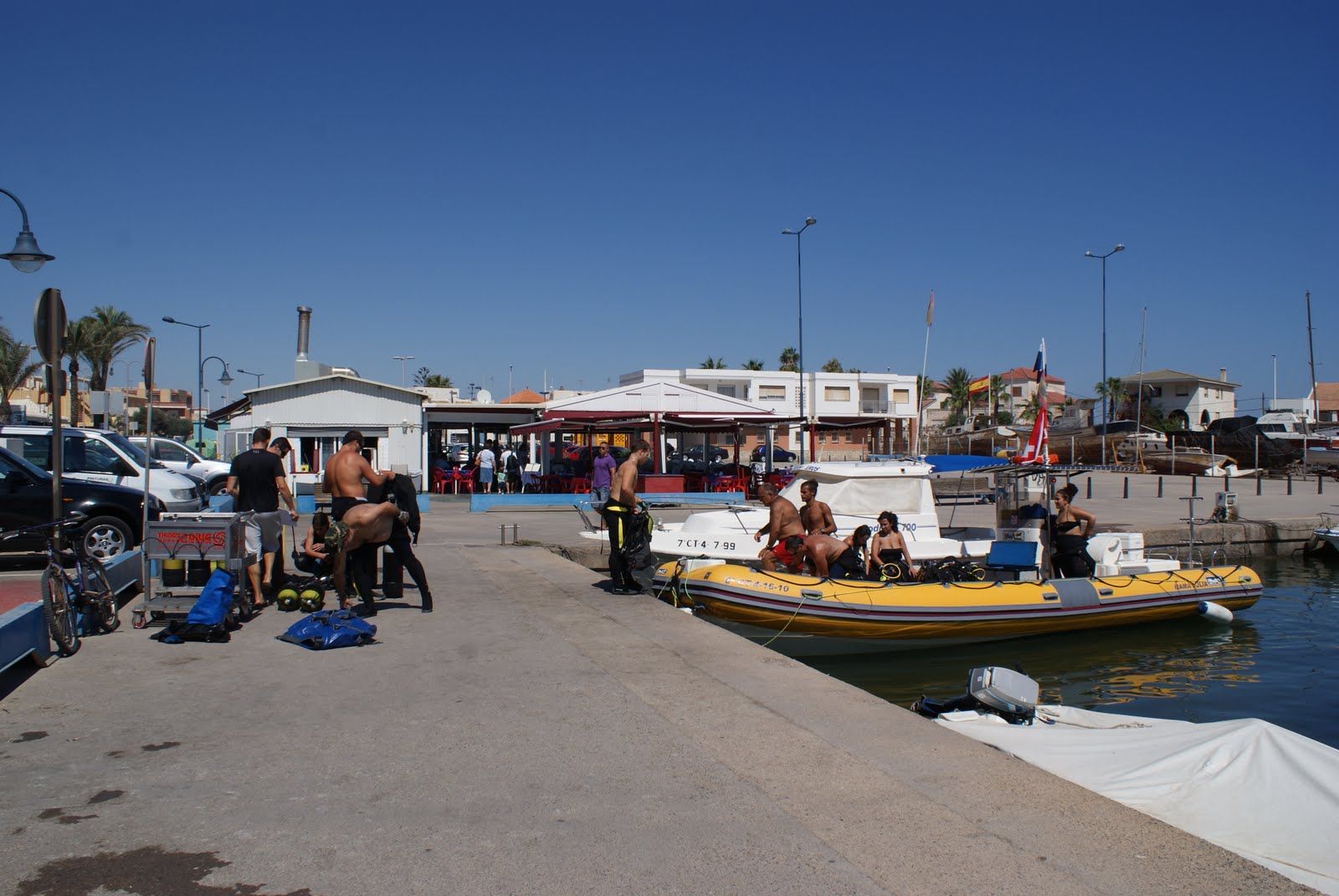 Escena del muelle con buzos preparándose para bucear, barco amarillo, cielo azul y edificios al fondo.