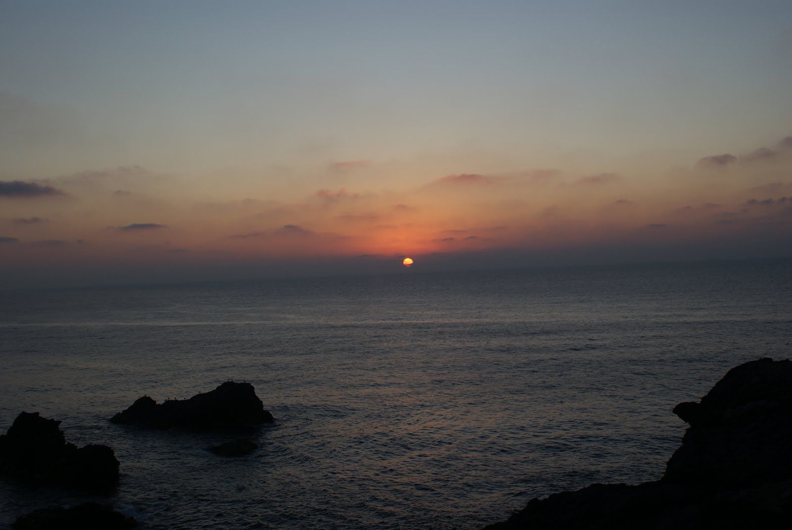 Puesta de sol sobre el océano, sol naranja cerca del horizonte; silueta oscura de rocas en primer plano.