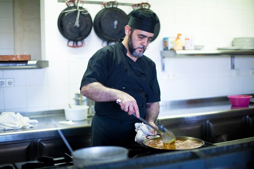 Un chef con uniforme negro cocina en la estufa de una cocina comercial, revolviendo la comida en una sartén grande.
