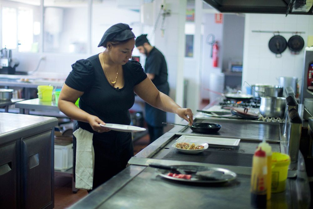 Mujer en uniforme negro cocinando en una cocina comercial, con platos en el mostrador y el chef al fondo.