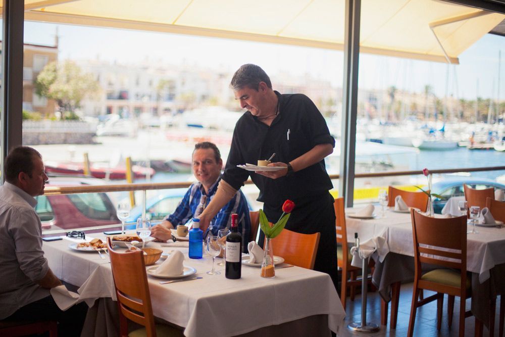 Camarero sirviendo un plato a un hombre sentado en una mesa en un restaurante frente al mar.