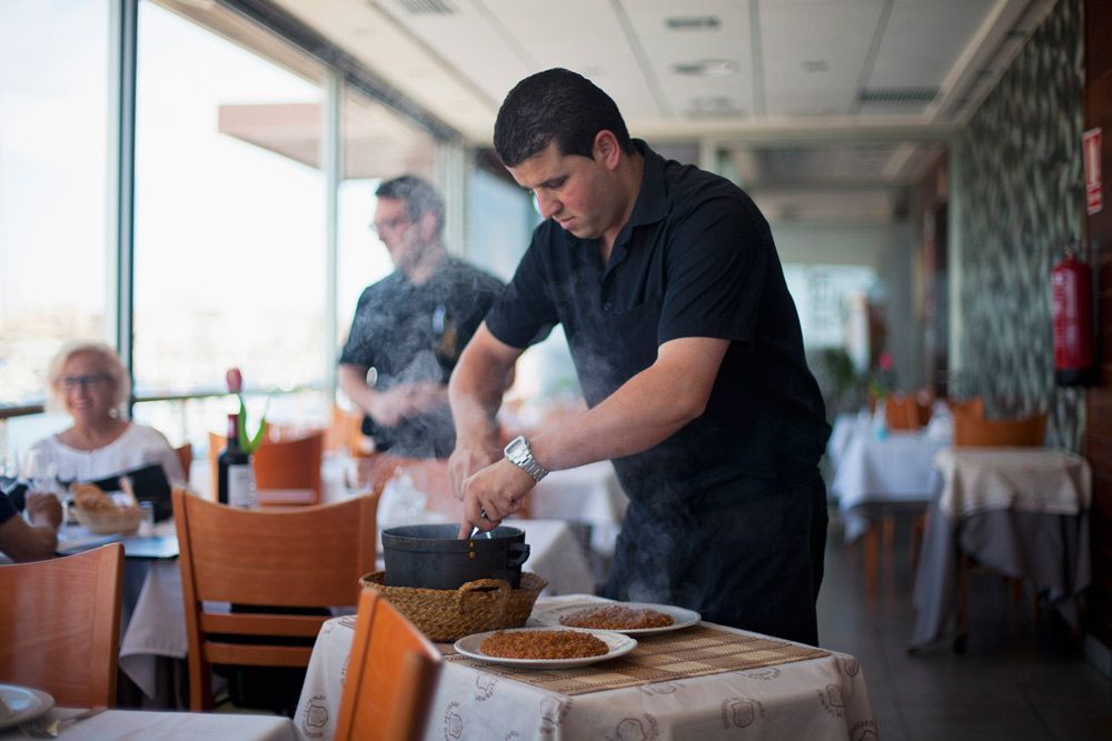 Chef sazonando comida en un restaurante, un camarero al fondo, un cliente sentado.