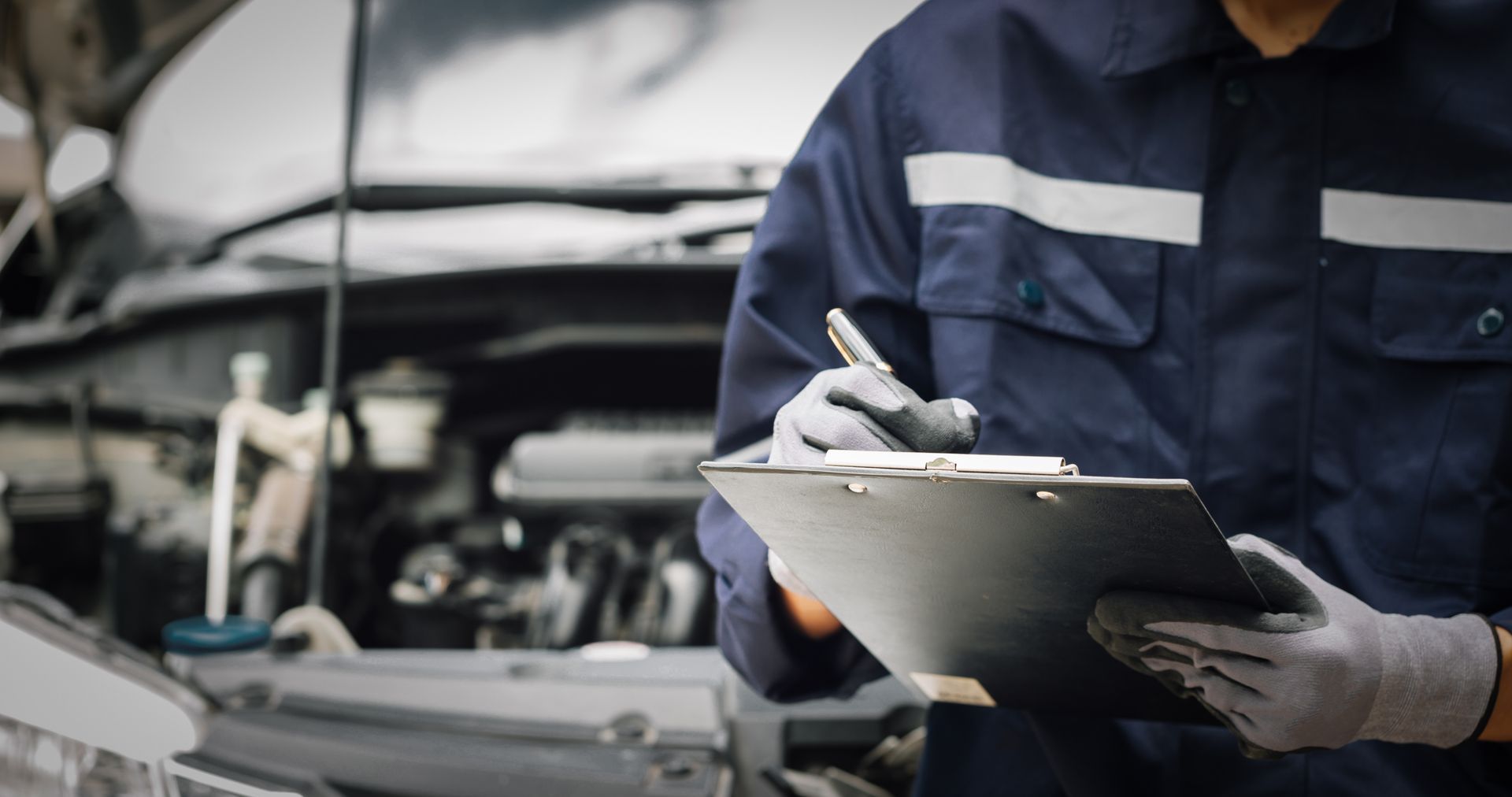 Un mécanicien en combinaison bleue prend des notes sur un bloc-notes à côté du capot ouvert d'une voiture dans un garage.