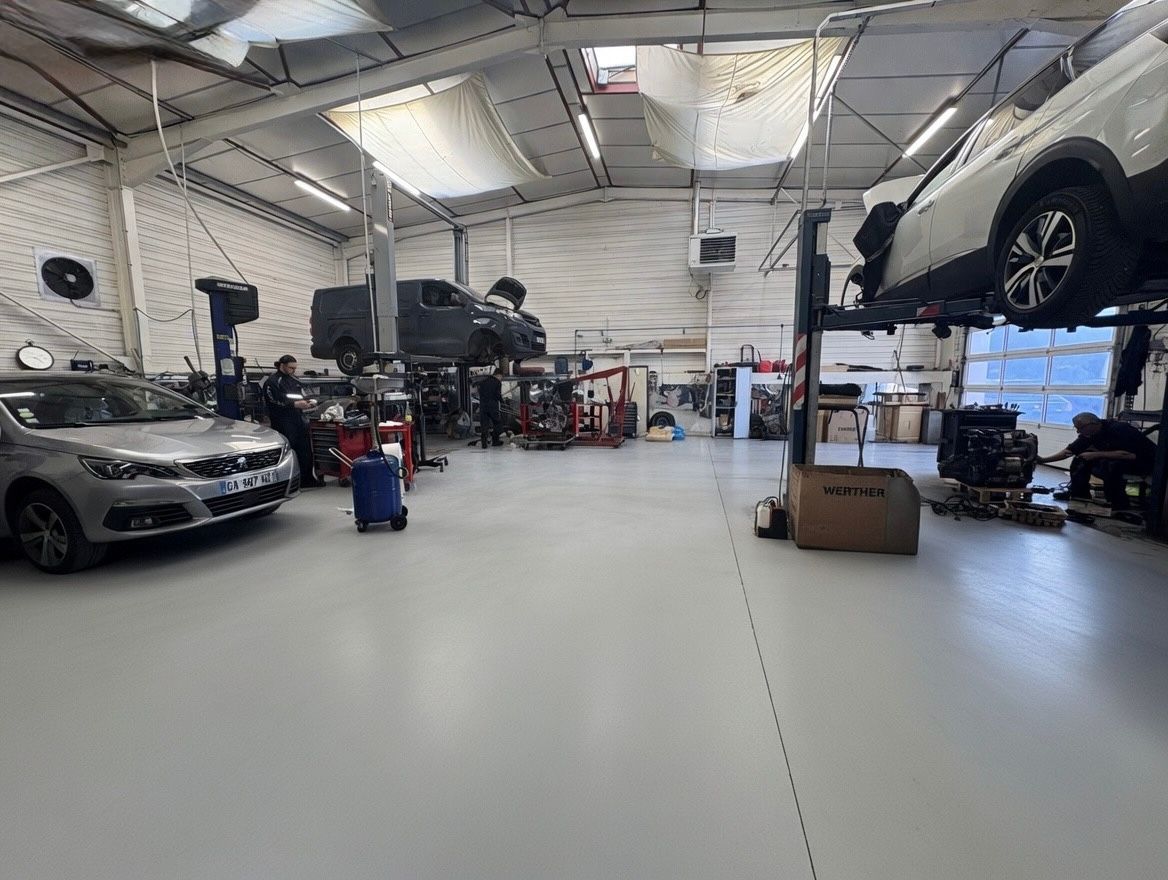 Atelier de réparation automobile avec des voitures sur des ponts élévateurs, une voiture argentée à gauche et des cartons sur un sol blanc propre.