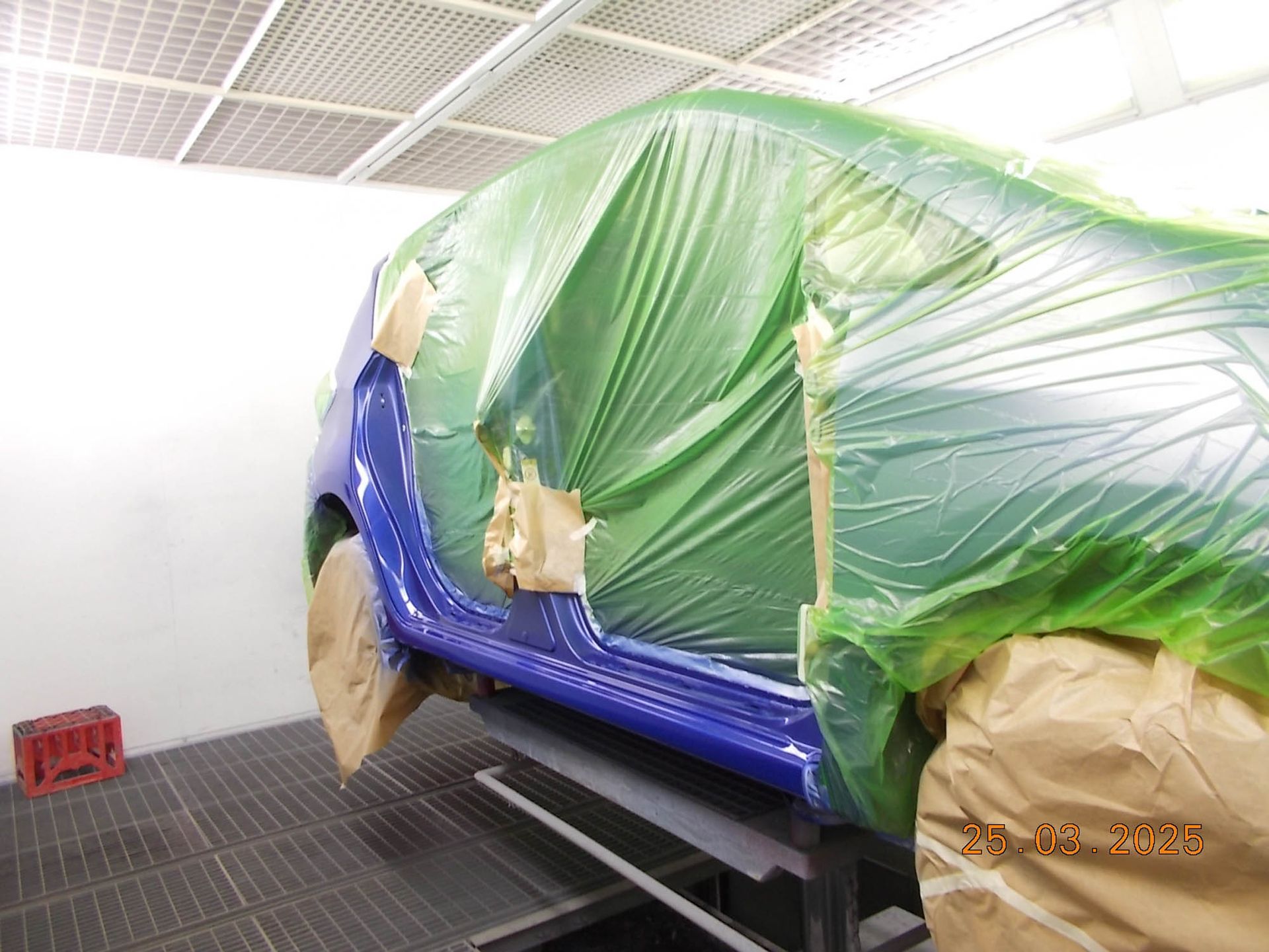 In einer Lackierkabine wird ein blaues Auto mit grüner Plastikfolie abgedeckt.