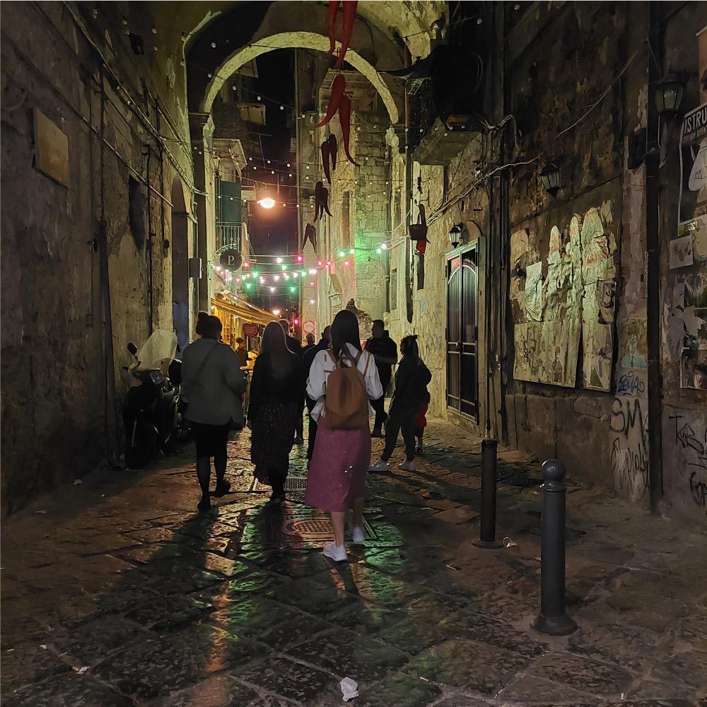 Gente caminando por un callejón estrecho y poco iluminado con una entrada arqueada y luces festivas.