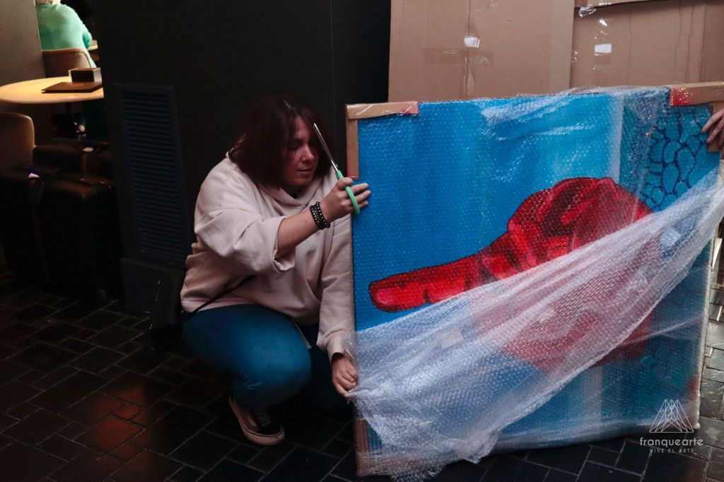 Mujer cortando plástico de burbujas de un cuadro grande. La obra tiene una mano roja sobre fondo azul. Interior oscuro.
