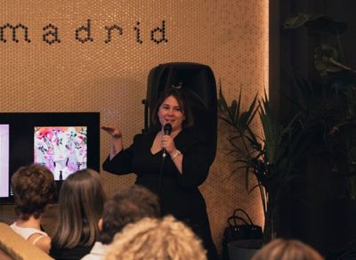 Una mujer habla por un micrófono y gesticula durante una presentación en un local de Madrid. El público escucha.