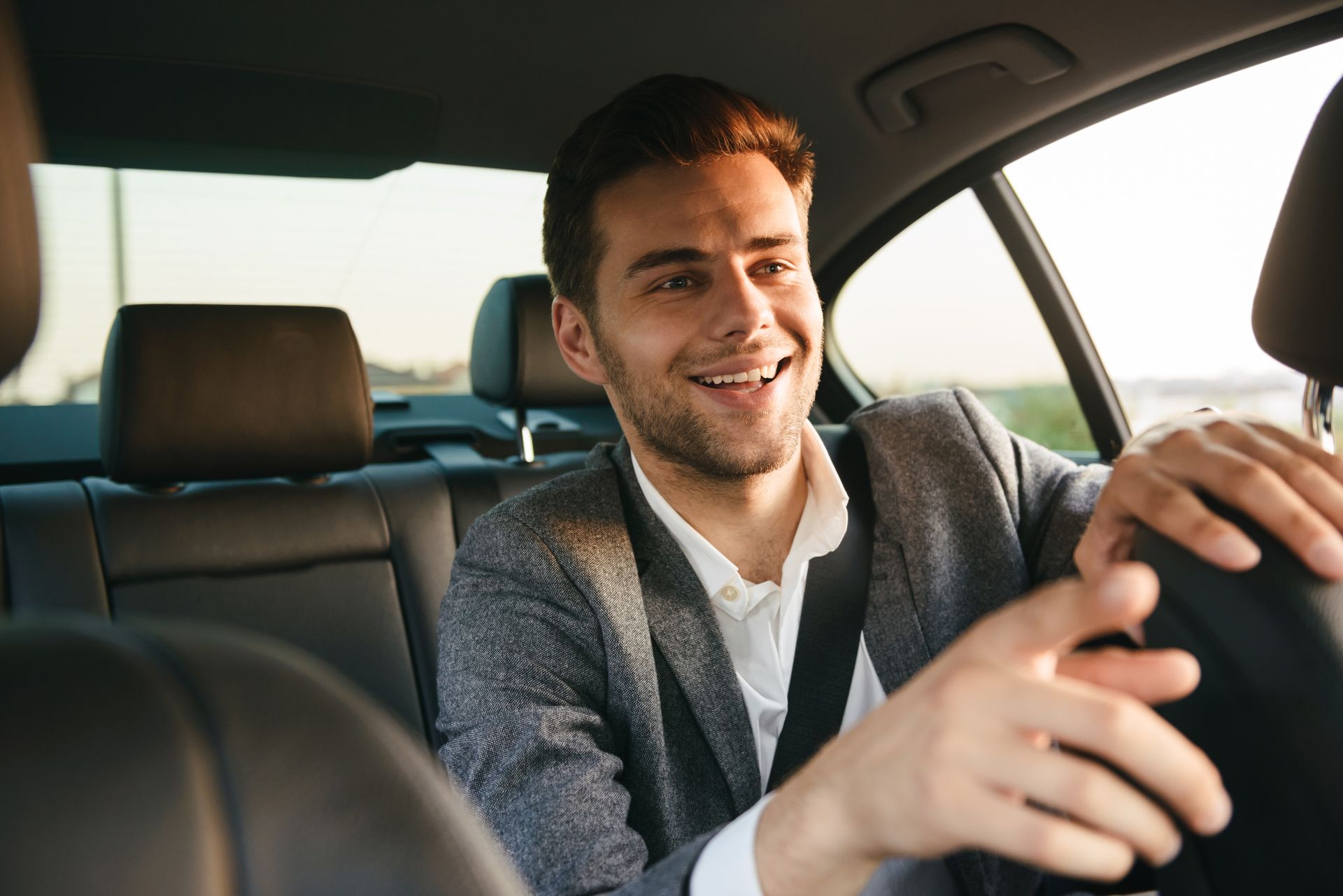 Un conducteur souriant, vêtu d'un blazer, conduit une voiture, vu du siège avant.