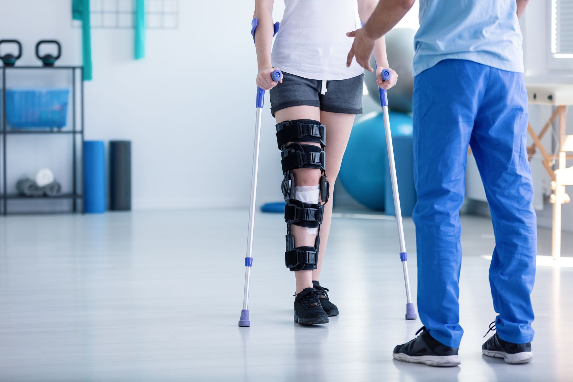 Un physiothérapeute aide un patient portant une attelle à la jambe à se déplacer avec des béquilles dans une salle de consultation lumineuse.