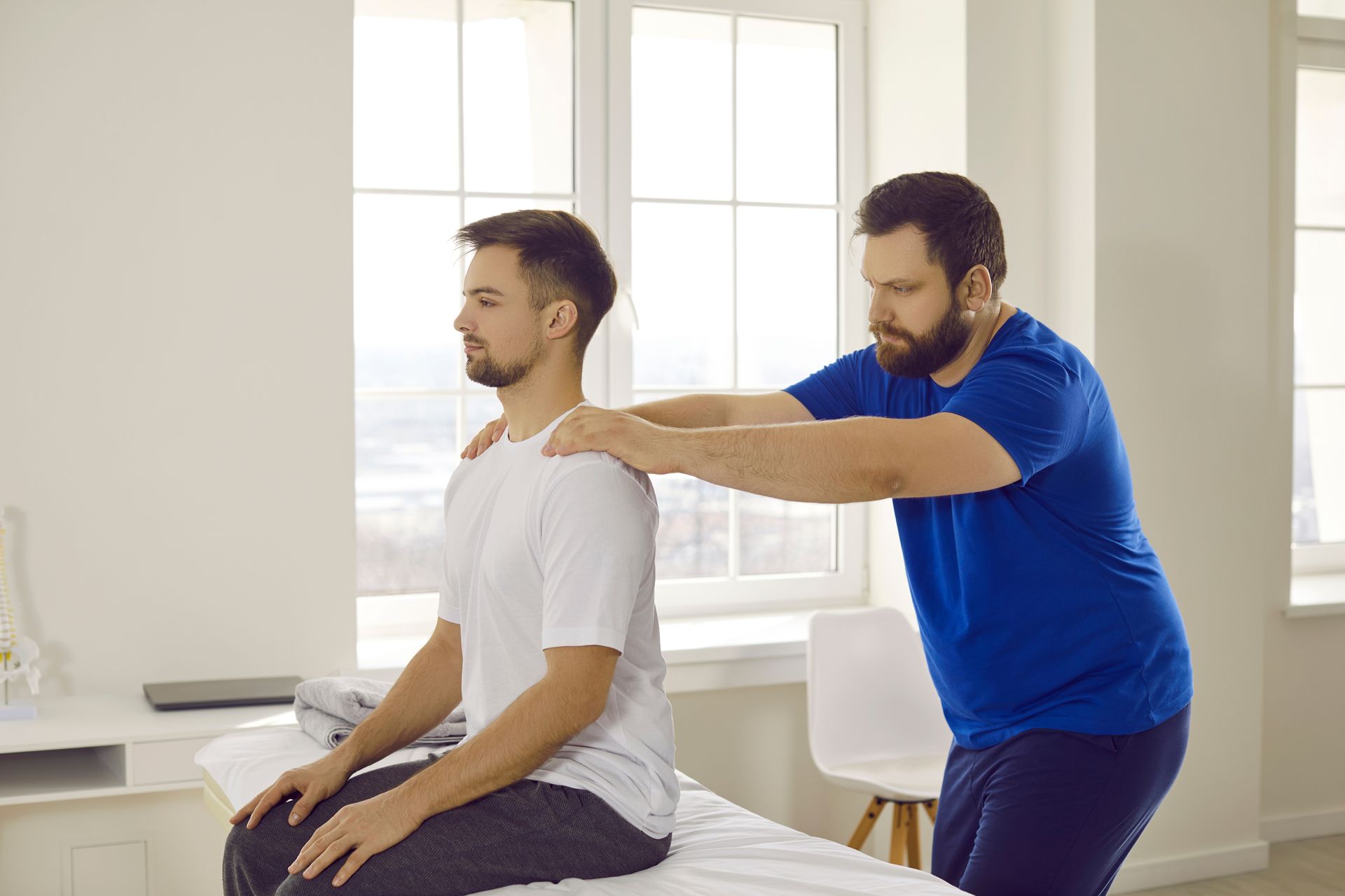 Un physiothérapeute ajuste les épaules d'un homme assis dans une salle de clinique lumineuse.