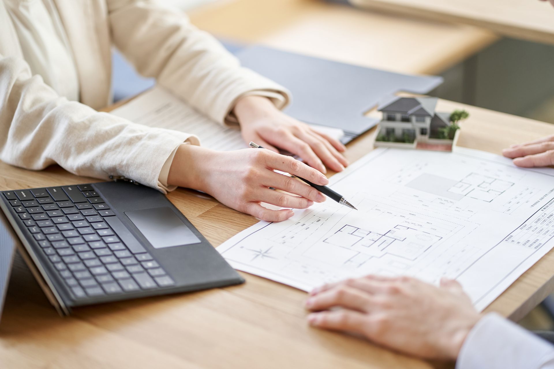 Manos señalando planos sobre una mesa junto a un ordenador portátil y una maqueta de casa.