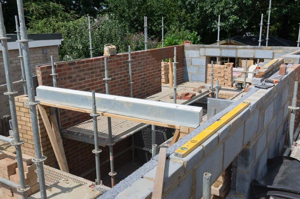 Un chantier de construction montrant des murs en briques rouges et en parpaings gris à moitié construits, avec des échafaudages et un niveau à bulle jaune.