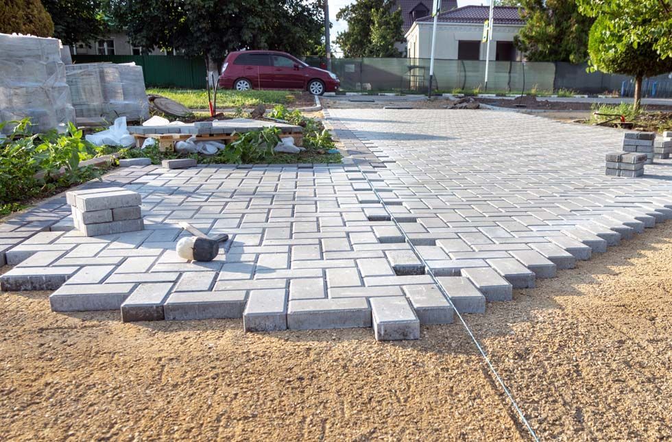 Allée carrossable partiellement achevée, composée de dalles rectangulaires grises posées en chevrons sur une base de gravier.