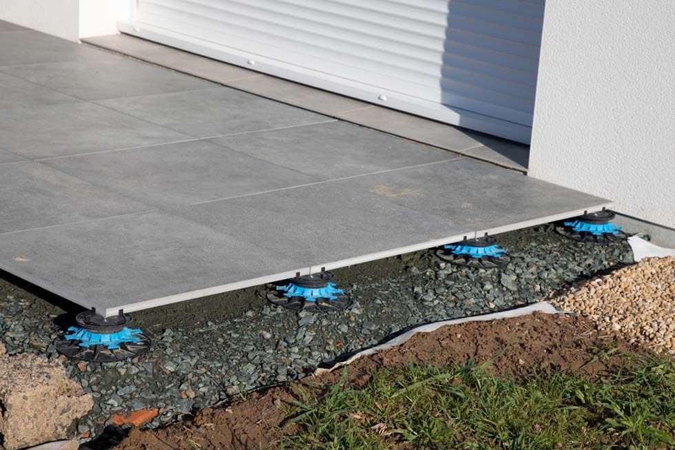 Des carreaux d'extérieur gris reposent sur des socles réglables en plastique bleu, au-dessus d'un lit de gravier, contre un mur de bâtiment.