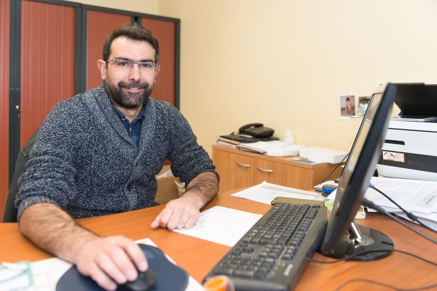 Un homme portant un pull gris et des lunettes est assis à un bureau et utilise une souris d'ordinateur.