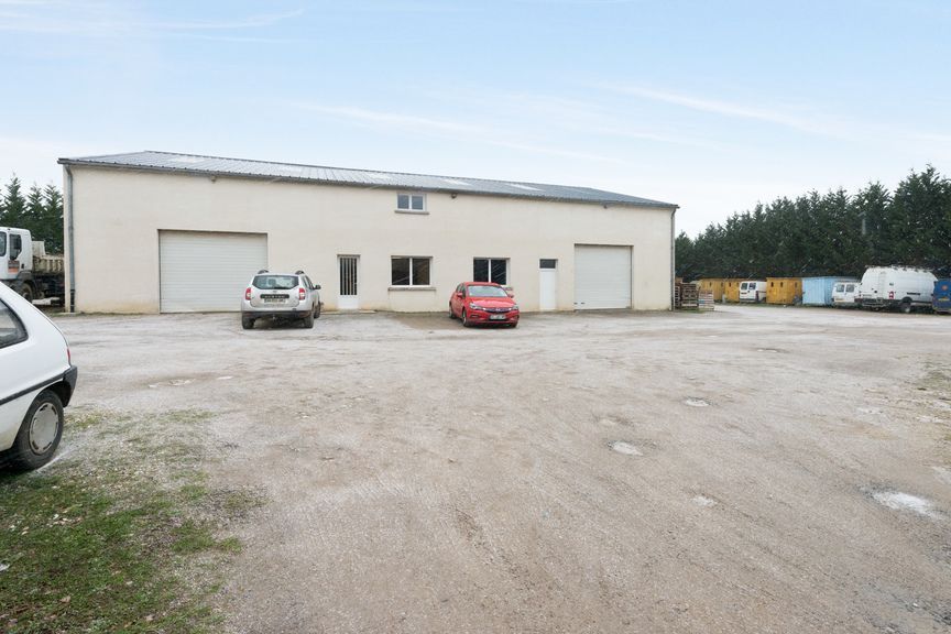 Un entrepôt beige avec deux portes de garage se dresse sur un terrain gravillonné, des voitures étant garées devant et des arbres se profilant au loin.