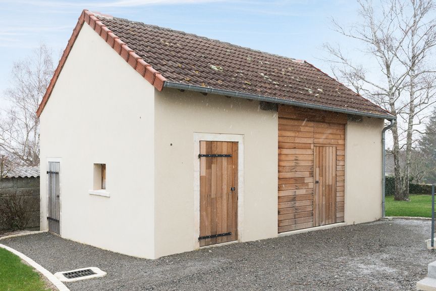 Un petit bâtiment de stockage couleur crème, avec un toit de tuiles brunes et deux portes en bois, entouré de gravier.