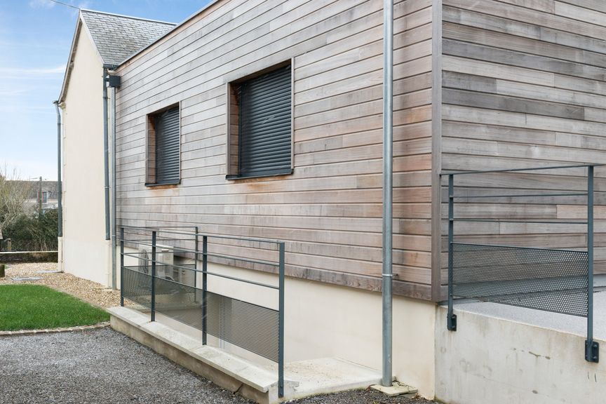 Vue latérale d'une maison moderne avec des murs lambrissés, une rambarde de sécurité en métal et une fenêtre de sous-sol au niveau du rez-de-chaussée.
