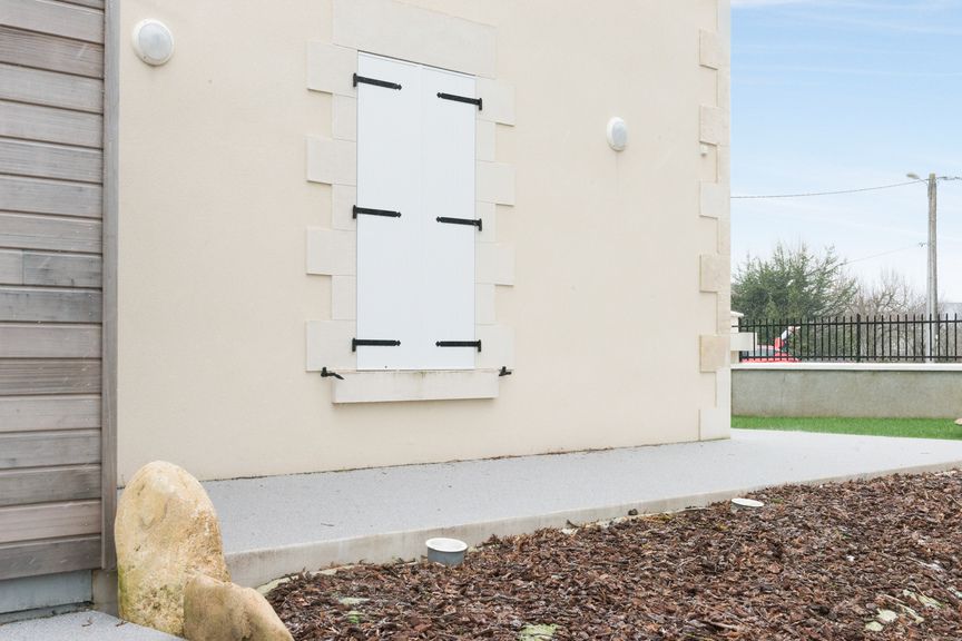Un mur beige avec des volets blancs, un sol clair et un tapis de paillis au premier plan.