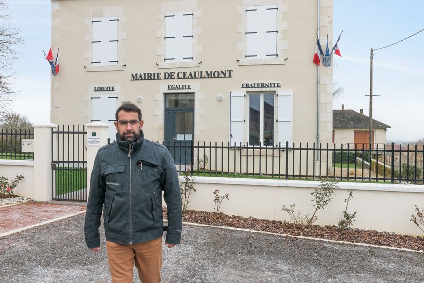 Une personne se tient devant la mairie de Ceaulmont, un bâtiment municipal de couleur claire avec des volets blancs et des drapeaux français.
