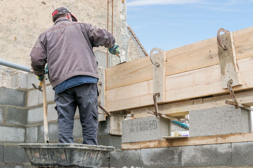 Un ouvrier du bâtiment en veste se tient sur un muret, en train d'installer un coffrage en bois pour une poutre en béton sur un chantier.