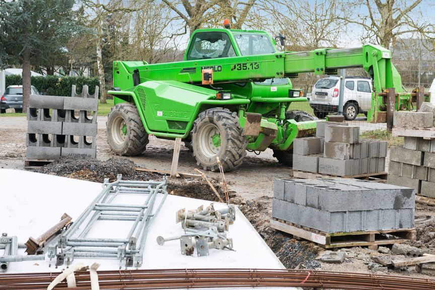 Un chariot télescopique Merlo vert vif est posé sur un terrain vague, entouré de piles de blocs de béton et de matériaux de construction.