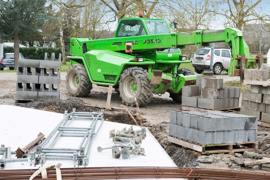 Un chariot télescopique vert vif est stationné sur un chantier de construction, à proximité de palettes de blocs de béton et d'échafaudages métalliques.