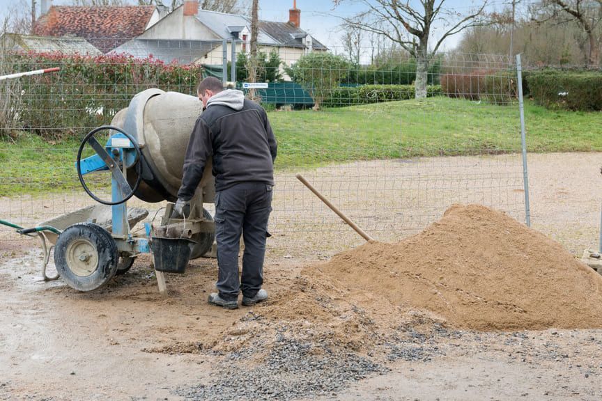 Un ouvrier vêtu d'une veste et d'un pantalon sombres manœuvre une bétonnière portative bleue près d'un tas de sable sur un chantier extérieur.