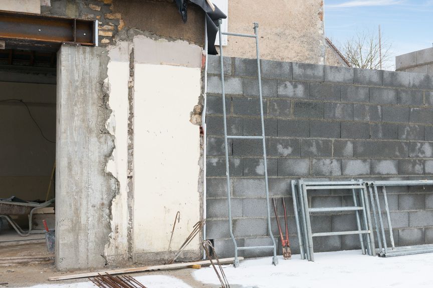 Un chantier de construction présentant un mur en béton inachevé et une porte avec un linteau en acier, à côté d'un amas d'échafaudages.