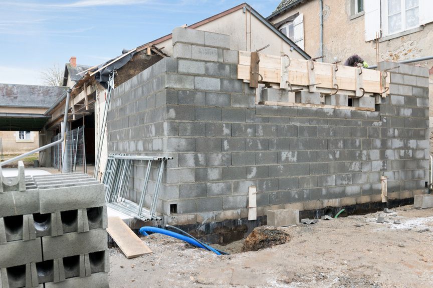Chantier de construction montrant un mur en parpaings gris en cours d'assemblage, des coffrages en bois pour une poutre en béton et une cour en terre.
