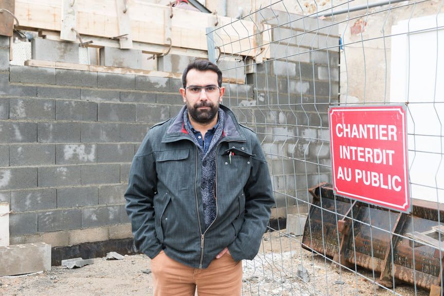 Un homme se tient devant un chantier de construction avec un panneau rouge sur lequel on peut lire CHANTIER INTERDIT AU PUBLIC.
