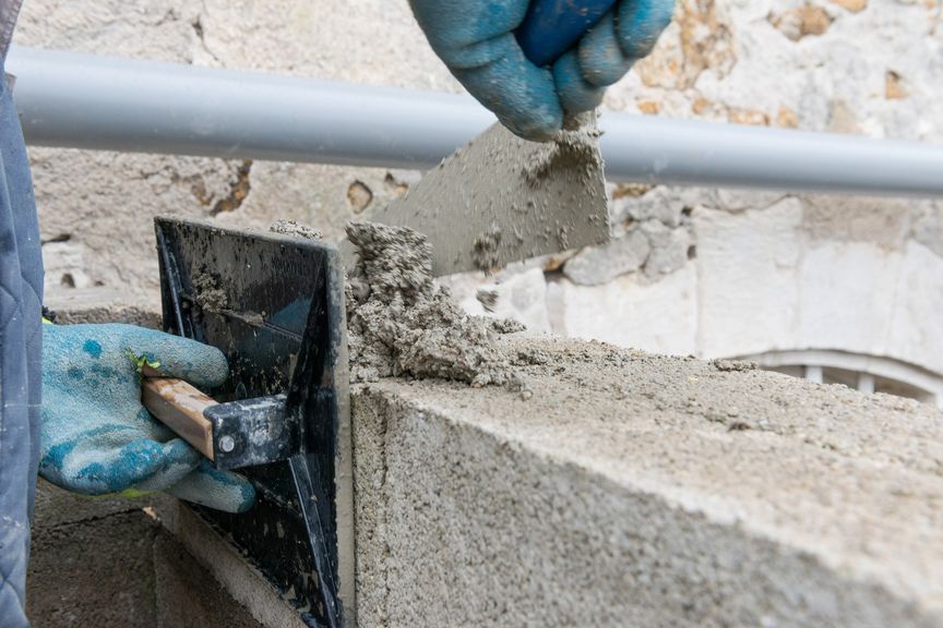 Un ouvrier portant des gants bleus utilise une truelle pour appliquer du mortier sur un mur en blocs de béton.
