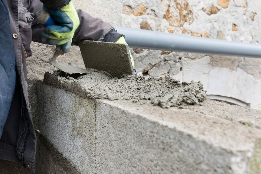 Une main gantée utilise une truelle métallique pour étaler du mortier gris sur le dessus d'un bloc de béton brut.