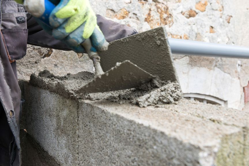Une main gantée utilise une truelle métallique pour étaler du mortier sur un bloc de béton gris pendant la construction.
