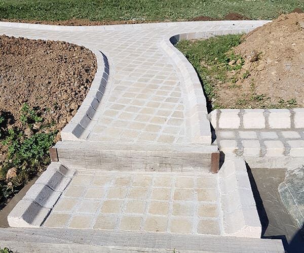 Une allée pavée avec une marche peu profonde, des dalles de pierre claire et des bordures en béton dans un cadre de jardin.