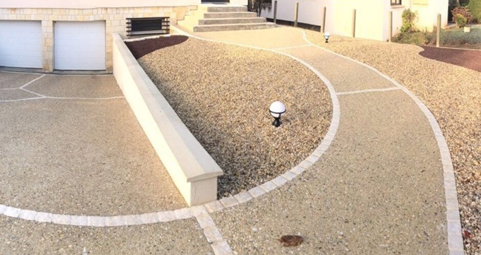 Une allée et un chemin d'accès pavés de gravier beige, délimités par des bordures en pierre claire, mènent à l'entrée de la maison.