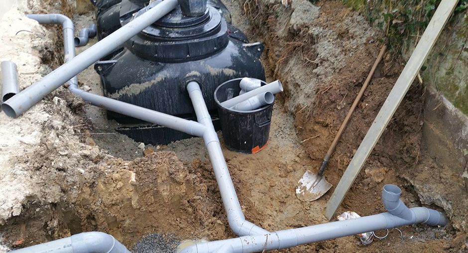 Une fosse septique en plastique noir est installée dans une tranchée de terre avec des tuyaux de drainage en PVC gris et une pelle.